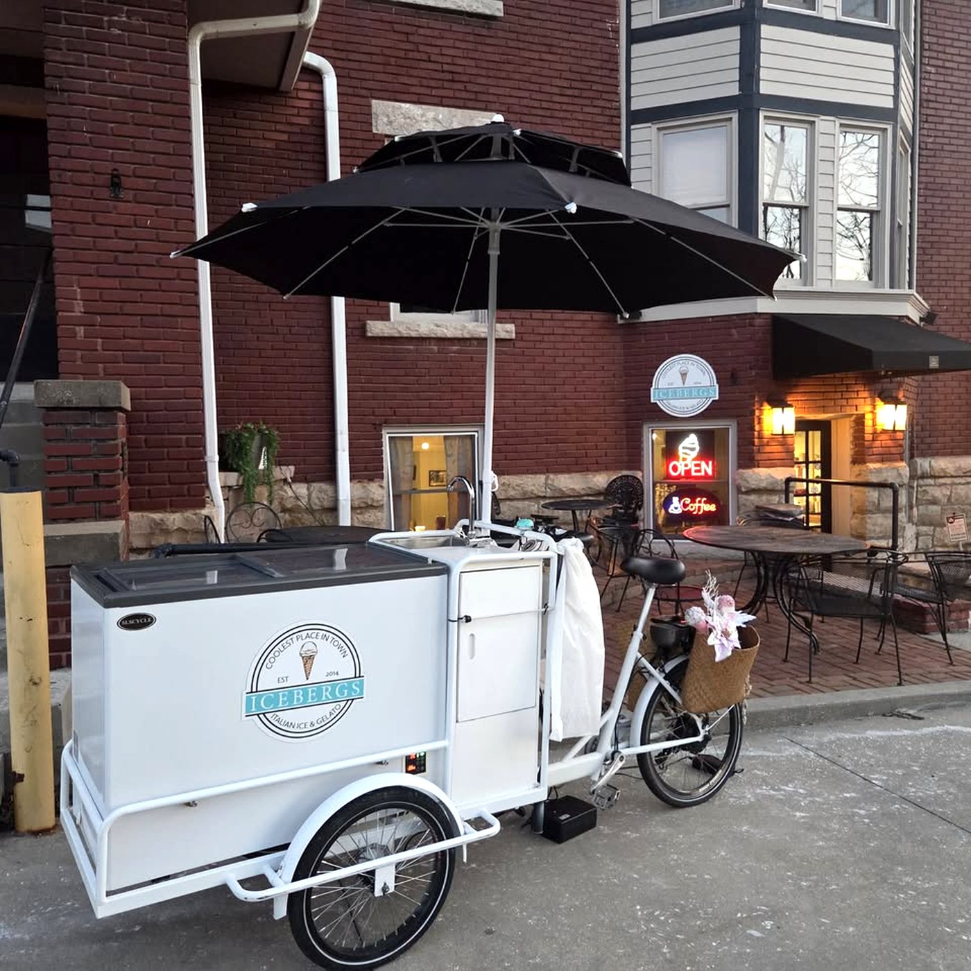 The white Icebergs gelato tricycle with its black umbrella parked outside the shop on Water Street at dusk, the neon Open sign lit behind it.
